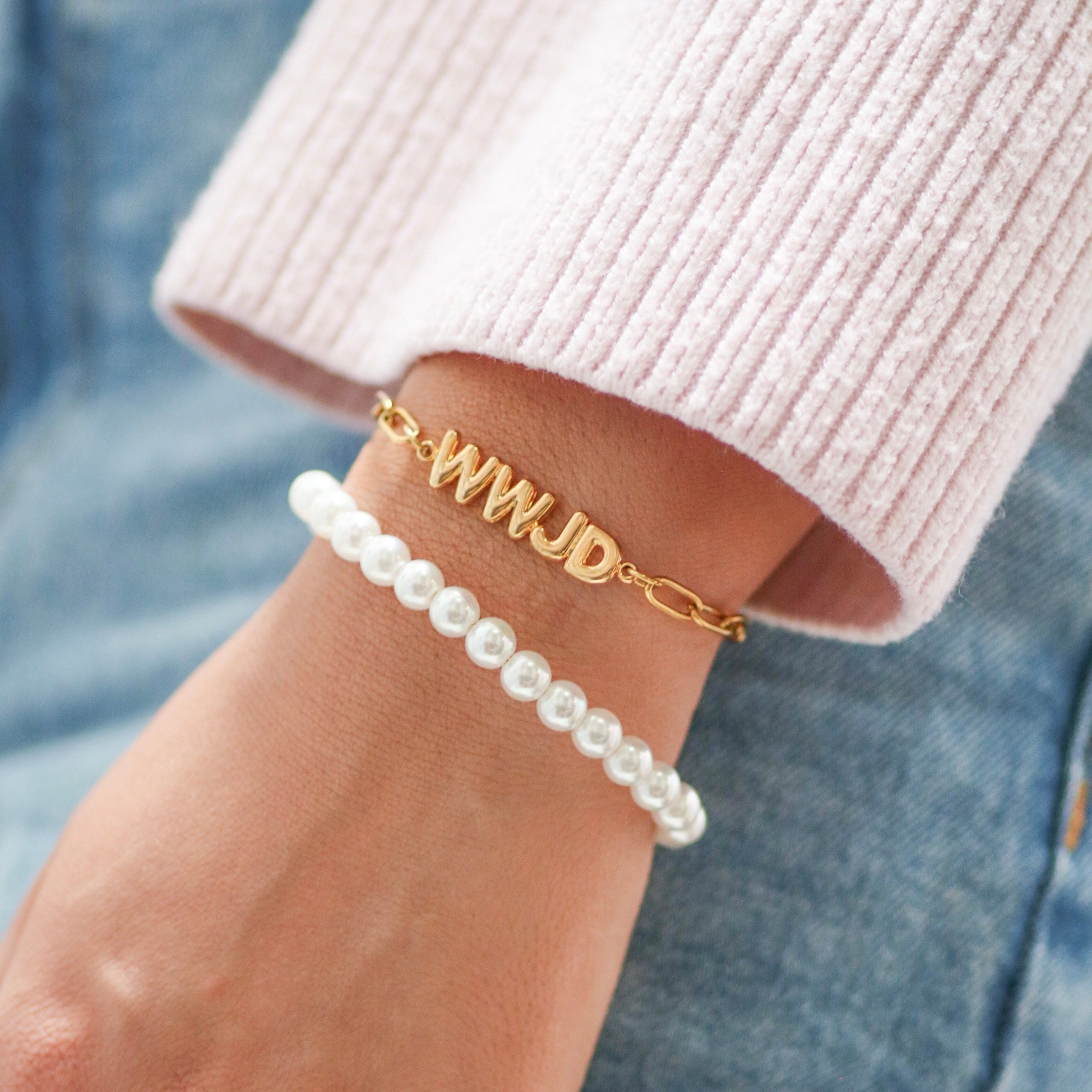 Hand wearing a gold bracelet with 'WWJD' and a pearl bracelet on a denim background